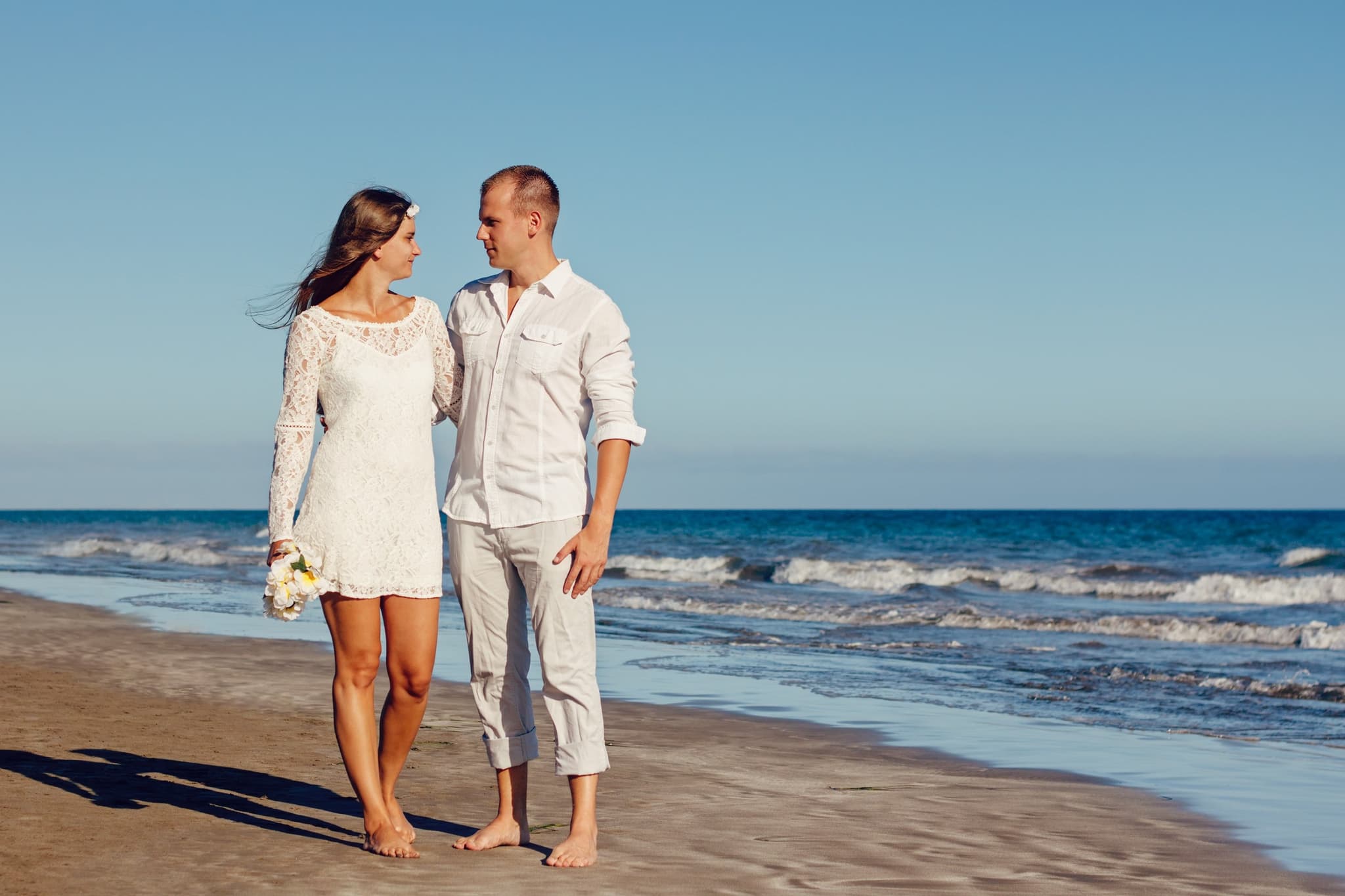 A couple walking barefoot along the beach in white, soft sunlight and a blue sea